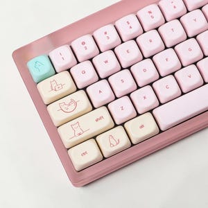 May include: A pink mechanical keyboard with light pink keycaps. The keycaps feature cat illustrations and text. The keyboard has a light blue escape key with a cat face. The keyboard is angled, showing the keys and the pink frame.