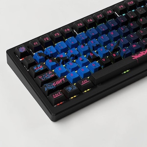 May include: A black mechanical keyboard featuring blue keycaps with black paint splatter patterns and pink lettering. The shift key displays a skull and crossbones graphic, while other keys feature unique hand-drawn style illustrations.