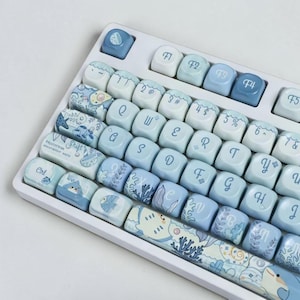 May include: A mechanical keyboard with a white frame and light blue keycaps featuring an ocean theme. The keys display letters, numbers, and function labels in a cursive font, alongside illustrations of sea creatures, coral, and waves.