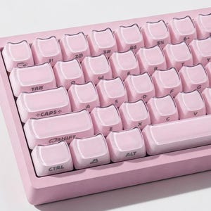 May include: A pink mechanical keyboard with cat-shaped keycaps. The keyboard has a light pink frame and keycaps with white lettering. The keys include letters, numbers, and special function keys like "TAB", "CAPS", "SHIFT", "CTRL", and "ALT".