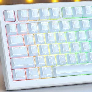 May include: A white mechanical keyboard with backlit keys. The keys are white with black lettering, and the backlighting transitions through a rainbow of colors. The keyboard has a minimalist design and a rectangular shape.