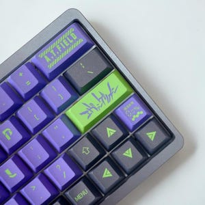 May include: A close-up of a mechanical keyboard with purple and gray keys. The keyboard has a gray frame and features green and purple accent keys with text including "A.T.FIELD" and Japanese characters. The keycaps are a mix of purple and gray.