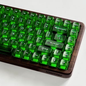 May include: A mechanical keyboard featuring translucent emerald green keycaps with white lettering set into a dark brown wooden frame. The Womier brand name is printed in black on the lower right side of the wooden base, which includes standard function, navigation, and alphanumeric keys.