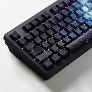 May include: A mechanical keyboard featuring a black frame and keycaps with a dark galaxy print. The keys display white letters and symbols, with subtle RGB backlighting visible between the keys. The design includes a space-themed aesthetic.