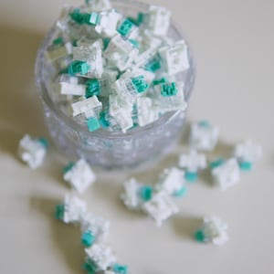 May include: A collection of mechanical keyboard switches featuring white housings and teal stems. Many of these small, square-shaped components are contained within a clear glass bowl, while several others are scattered around the base.