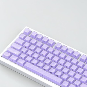 May include: A mechanical keyboard featuring a white frame and light purple keycaps. Each keycap displays a subtle diagonal textured pattern and standard black QWERTY lettering. The layout includes function keys, a space bar, and modifier keys.