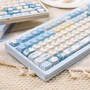 May include: A light blue mechanical keyboard with white and light blue keycaps. The keycaps feature various designs, including clouds and cupcakes. The keyboard has a light blue frame and a white base. The keyboard is a standard size.