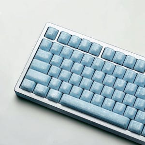 May include: A mechanical keyboard featuring a silver frame and light blue keycaps with a marbled, translucent texture. The keys are blank, lacking any printed letters or symbols, and are arranged in a standard layout within the metallic casing.