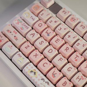 May include: A white keyboard with custom pink keycaps. The keycaps feature cat-themed designs and pastel colors. The keys display letters, numbers, and symbols, with some keys labeled "Tab", "Caps", and "Alt".