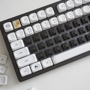 May include: A black mechanical keyboard with white keycaps. Some keycaps have custom designs, including a cat and a wizard. The keyboard is on a white surface, with several loose keycaps scattered around.