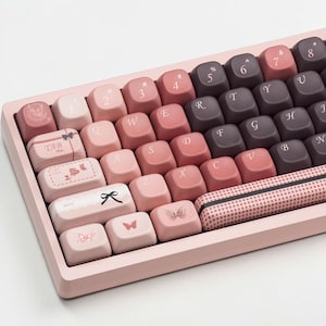 May include: A pink mechanical keyboard with a gradient color scheme. The keycaps feature a mix of pink and dark purple hues, with decorative designs and text. The keyboard has a rectangular shape and a matching pink frame.