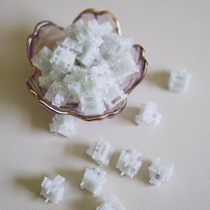 May include: A collection of translucent white mechanical keyboard switches, with several contained in a small, scalloped, purple-tinted glass dish with a gold rim, and others scattered around the base of the dish on a neutral surface.