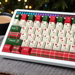 May include: A white mechanical keyboard featuring custom holiday-themed keycaps. The keys are cream-colored with red and green accents, displaying festive icons like candy canes, holly, and bows. The spacebar and modifier keys feature a red plaid pattern.