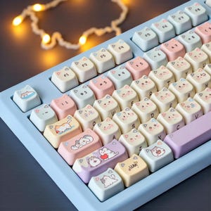 May include: A light blue mechanical keyboard with cat-shaped keycaps. The keycaps are a mix of pastel colors, each featuring a different cat illustration. The keyboard has a light blue frame and a lavender space bar.