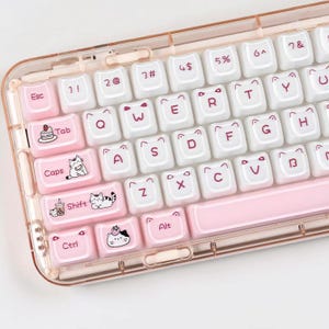 May include: A mechanical keyboard with a translucent peach-colored frame and white keycaps. The keys feature dark pink lettering, cat-ear accents, and illustrations of cats, strawberries, and boba tea on the Esc, Tab, Caps, Shift, and Ctrl keys.