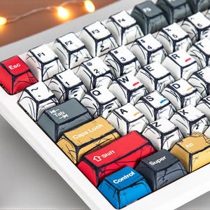 May include: A white mechanical keyboard with custom keycaps. The keycaps feature a unique black and white sketch design with colorful accents on the modifier keys. The "Esc" key is red, and the "Shift" key is red.