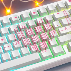 May include: A white mechanical keyboard with custom keycaps. Some keycaps feature pink heart symbols and pink striped designs. The keyboard has a green and blue underglow.