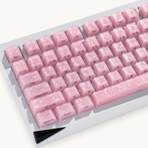 May include: A computer keyboard featuring a white frame and translucent pink keycaps with a marbled, cloudy white texture. The keys display standard QWERTY layout characters, including function keys, letters, numbers, and symbols, with a black accent piece.
