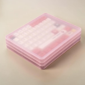 May include: A rectangular, semi-transparent pink plastic storage case containing a collection of small, square mechanical keyboard keycaps in various shades of white, red, and dark gray, visible through the frosted lid.