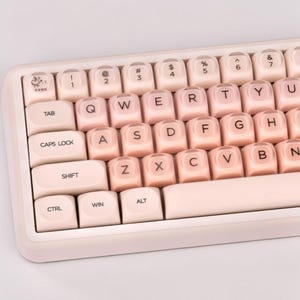 May include: A mechanical keyboard featuring soft pastel pink keycaps with rounded edges and dark brown lettering. The top left key displays a small graphic of a cartoon apple. The keyboard frame is a matching light pink color with a clean layout.