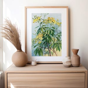 Op de afbeelding: Een ingelijste botanische prent met een aquarel illustratie van een bloeiende plant met gele bloemen en groene bladeren. Het kunstwerk wordt tentoongesteld op een houten dressoir, samen met decoratieve vazen en een klein kopje.
