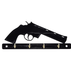 May include: Black key holder in the shape of a revolver. The key holder has a horizontal bar with four gold-colored hooks. The revolver silhouette is above the bar, with a trigger and barrel detail.
