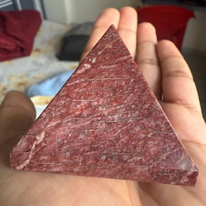 May include: A red jasper pyramid with a textured surface and white veining. The pyramid is held in a hand, showcasing its triangular shape and natural stone appearance. This decorative item is suitable for home decor or as a unique gift.