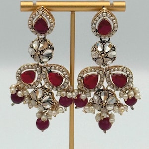 Peut inclure: Boucles d'oreilles dorées ornées de pierres rouge rubis et de petites perles. Les boucles d'oreilles présentent une pierre rouge rubis en forme de goutte d'eau en haut, avec d'autres pierres rouges rubis et des grappes de perles.