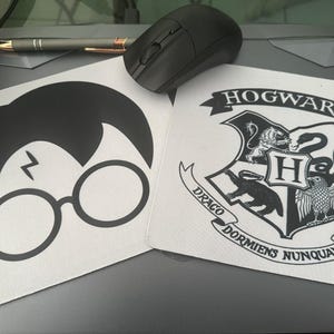 May include: Two white mouse pads with black graphics. One features a silhouette of Harry Potter's hair, glasses, and lightning bolt scar. The other displays the Hogwarts crest with the words "HOGWARTS" and "DRACO DORMIENS NUNQUAM TITILLANDUS". A black mouse and pen are on top.
