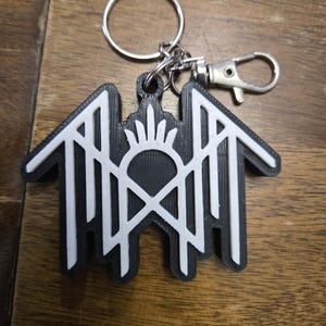 May include: A black and white keychain featuring a stylized geometric emblem with a central sunburst motif and symmetrical angular lines. The design is constructed with a layered appearance, attached to a silver-toned metal split ring and a lobster clasp.