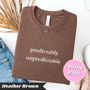 May include: A heather brown t-shirt with the words "predictably unpredictable" in white. A silver necklace with a sunburst pendant rests on the shirt. The shirt is folded, with a pair of blue jeans and a white shoe visible.