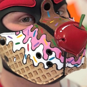 Peut inclure: Une cagoule en cuir noir avec un museau con&ccedil;u pour ressembler &agrave; un cornet de glace. Le museau pr&eacute;sente une base &agrave; texture gaufr&eacute;e, des motifs de gla&ccedil;age rose et blanc avec des vermicelles color&eacute;s et une grosse cerise rouge brillante.