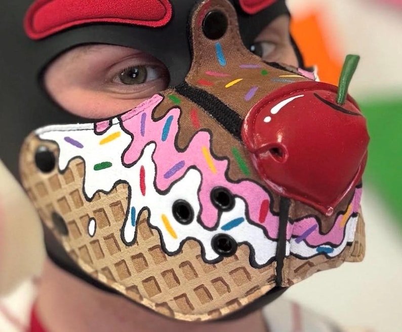 Peut inclure: Une cagoule en cuir noir avec un museau con&ccedil;u pour ressembler &agrave; un cornet de glace. Le museau pr&eacute;sente une base &agrave; texture gaufr&eacute;e, des motifs de gla&ccedil;age rose et blanc avec des vermicelles color&eacute;s et une grosse cerise rouge brillante.