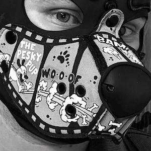 Peut inclure: Un masque animalier noir et blanc avec un imprim&eacute; style bande dessin&eacute;e portant les inscriptions THE PESKY PUP, WO-O-OF et BAM. Le masque comprend un embout nasal arrondi pro&eacute;minent, plusieurs &oelig;illets de ventilation circulaires et un motif graphique repr&eacute;sentant un chien qui court, des empreintes de pattes et des os.