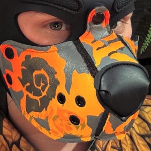 Peut inclure: Un masque museau avec un motif camouflage orange et gris vif. Le masque comprend une truffe noire pro&eacute;minente, des &oelig;illets m&eacute;talliques noirs pour la ventilation et des boutons-pression noirs sur les bords, fix&eacute; sur une cagoule noire.