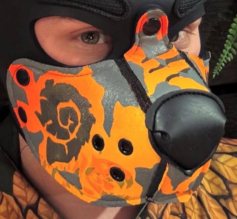 Peut inclure: Un masque museau avec un motif camouflage orange et gris vif. Le masque comprend une truffe noire pro&eacute;minente, des &oelig;illets m&eacute;talliques noirs pour la ventilation et des boutons-pression noirs sur les bords, fix&eacute; sur une cagoule noire.