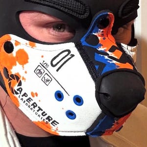 Peut inclure: Un masque museau de style animal blanc, orange et bleu avec le logo Aperture Laboratories, le num&eacute;ro 01 et de petites ic&ocirc;nes. Le masque pr&eacute;sente un embout nasal noir, des boutons-pression noirs et des &oelig;illets bleus sur une cagoule noire.