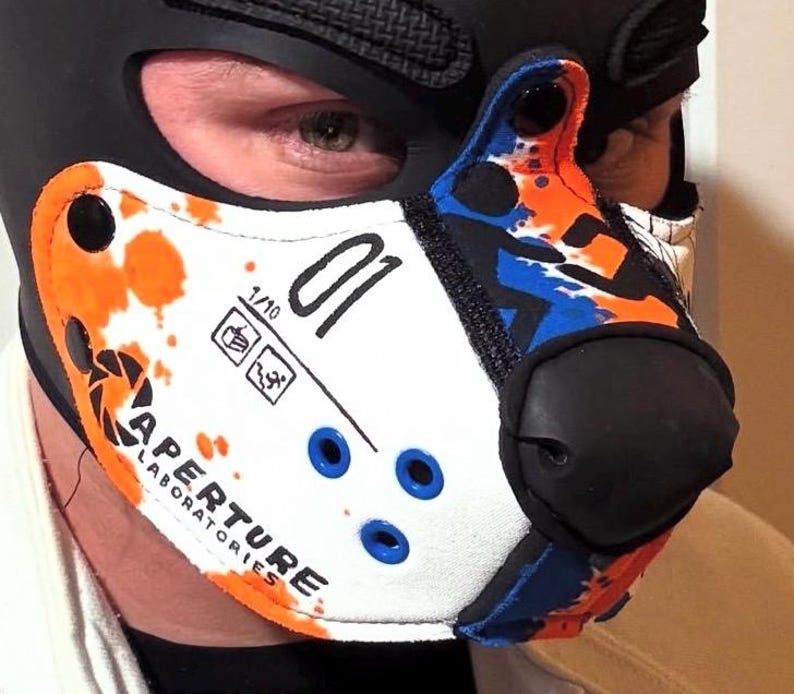 Peut inclure: Un masque museau de style animal blanc, orange et bleu avec le logo Aperture Laboratories, le num&eacute;ro 01 et de petites ic&ocirc;nes. Le masque pr&eacute;sente un embout nasal noir, des boutons-pression noirs et des &oelig;illets bleus sur une cagoule noire.