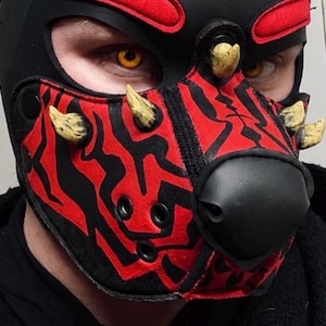 Peut inclure: Un masque cagoule &agrave; motifs noirs et rouges dot&eacute; d'un museau noir pro&eacute;minent, de plusieurs protub&eacute;rances beiges en forme de cornes et d'ouvertures pour les yeux. Le masque pr&eacute;sente un design rouge et noir dentel&eacute; sur le devant et le front.