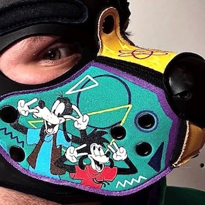Peut inclure: Une cagoule en similicuir noir avec un embout en forme de museau dot&eacute; d'un panneau turquoise. Le panneau pr&eacute;sente un graphique color&eacute; de deux chiens de dessin anim&eacute; faisant des signes de paix, avec des formes g&eacute;om&eacute;triques et des &oelig;illets.