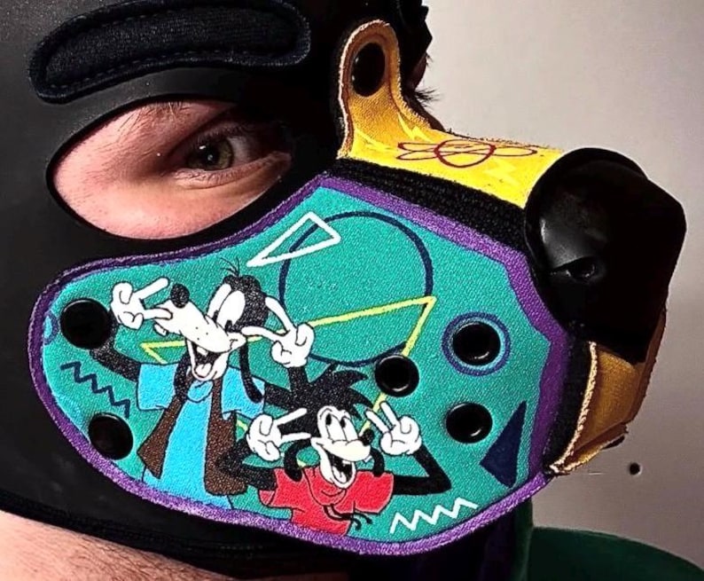 Peut inclure: Une cagoule en similicuir noir avec un embout en forme de museau dot&eacute; d'un panneau turquoise. Le panneau pr&eacute;sente un graphique color&eacute; de deux chiens de dessin anim&eacute; faisant des signes de paix, avec des formes g&eacute;om&eacute;triques et des &oelig;illets.