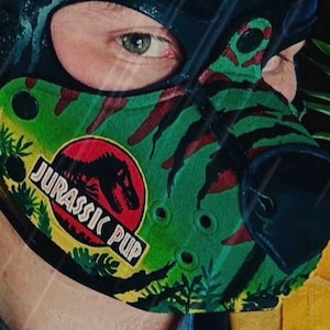 Peut inclure: Une cagoule noire avec une pi&egrave;ce de museau &agrave; motifs verts et rouges arborant un logo circulaire avec une silhouette de dinosaure noire et le texte JURASSIC PUP. Le museau pr&eacute;sente des accents noirs, des trous d'a&eacute;ration et un nez noir.