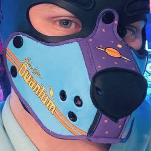 Peut inclure: Une cagoule noire avec un design &agrave; panneaux bleu clair et violet. La section bleu clair affiche le texte Nuka Cola Quantum en jaune avec des rayures orange. La section violette pr&eacute;sente des graphismes de plan&egrave;tes et d'&eacute;toiles orange.