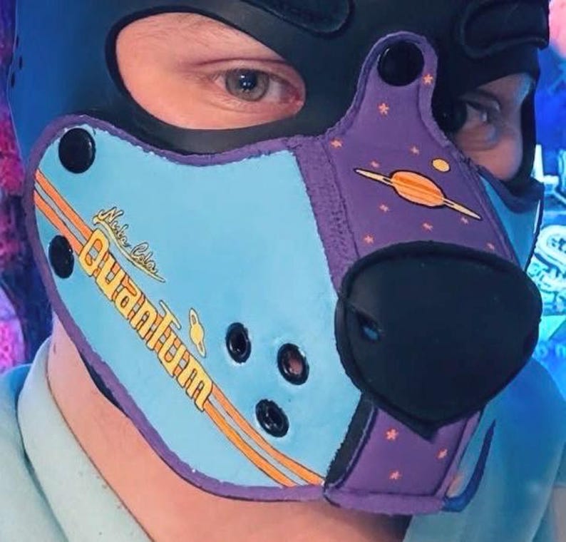 Peut inclure: Une cagoule noire avec un design &agrave; panneaux bleu clair et violet. La section bleu clair affiche le texte Nuka Cola Quantum en jaune avec des rayures orange. La section violette pr&eacute;sente des graphismes de plan&egrave;tes et d'&eacute;toiles orange.