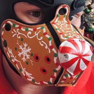 Peut inclure: Un masque facial beige arborant un motif de biscuit en pain d'&eacute;pices avec des d&eacute;corations de gla&ccedil;age blanc, des points rouges et verts, et un nez en forme de bonbon &agrave; la menthe ray&eacute; rouge et blanc. Il comporte des &oelig;illets m&eacute;talliques noirs.