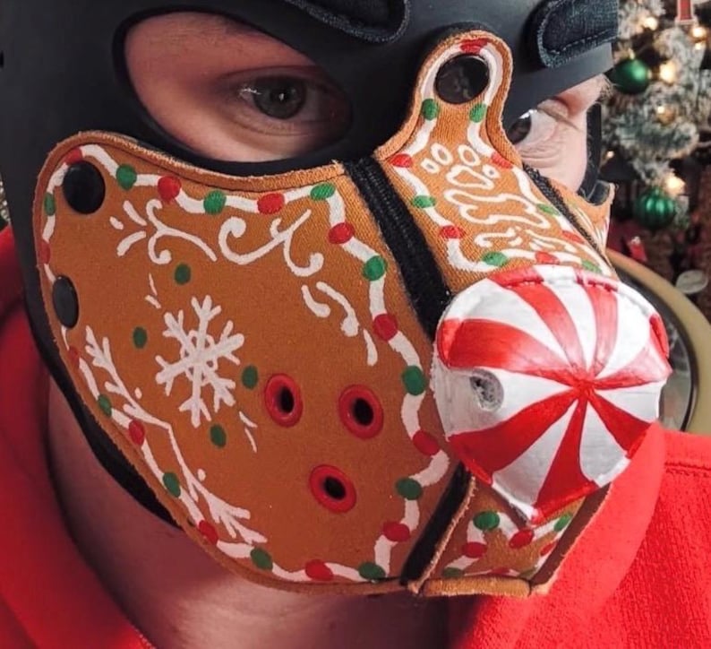 Peut inclure: Un masque facial beige arborant un motif de biscuit en pain d'&eacute;pices avec des d&eacute;corations de gla&ccedil;age blanc, des points rouges et verts, et un nez en forme de bonbon &agrave; la menthe ray&eacute; rouge et blanc. Il comporte des &oelig;illets m&eacute;talliques noirs.