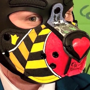 Peut inclure: Une cagoule noire dot&eacute;e d'un museau avec des rayures diagonales jaunes et noires, une section circulaire rouge et un nez en forme de c&oelig;ur. Le museau comprend des rivets noirs, un panneau sup&eacute;rieur d&eacute;coratif aux motifs gris et bleus et une bordure noire.