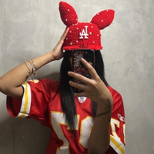 May include: A red crocheted hat with bunny ears, adorned with white pearls and the letter "A". The hat is worn with a red and white football jersey. The overall theme is a playful, handmade accessory.