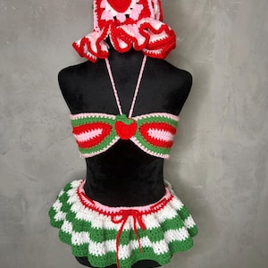 Puede incluir: Un conjunto de ganchillo hecho a mano que consta de un sombrero de pescador con temática de fresa, un top de bikini tipo halter con un motivo central de fresa en rojo, rosa y verde, y una falda con volantes y rayas horizontales blancas y verdes.