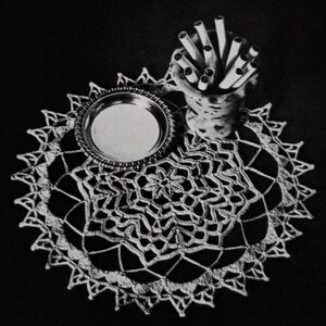 Könnte beinhalten: Eine weiße Häkeldeckchen mit einem zarten Blumenmuster, mit einem silbernen Tablett und einem Keramikhalter gefüllt mit gerollten Papier.